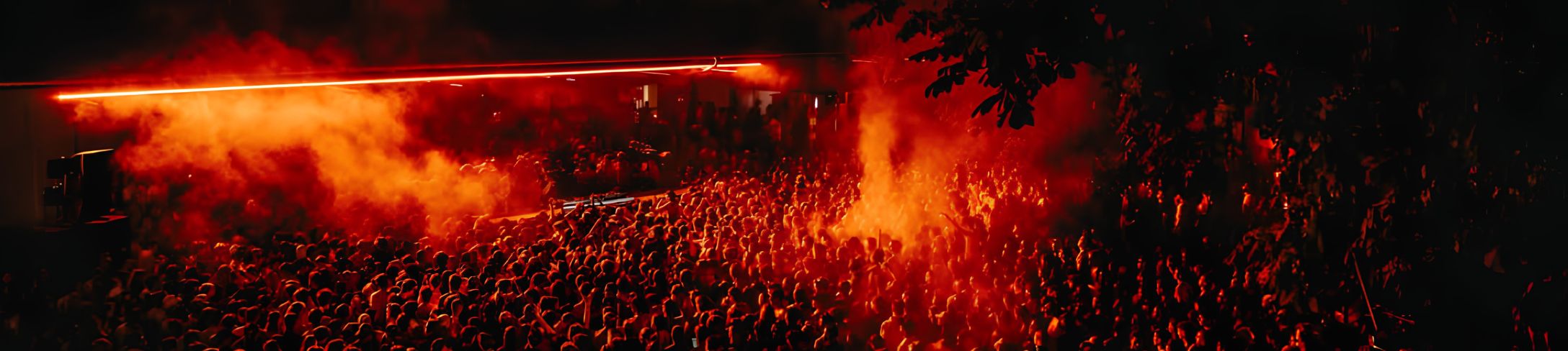 Totale eines Musikfestivals bei Nacht mit großem Publikum, das zu einem DJ-Pult schaut, rotes Licht und Dampf in der Luft