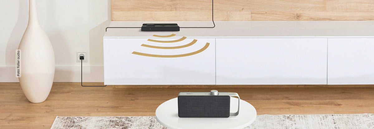 TV-Sprachverstärker OSKAR steht auf einem kleinen weißen Couchtisch vor einem Sideboard im Wonhzimmer, auf dem Sideboard ein TV-Receaver mit grafischen Signalwellen zum Sprachverstärker