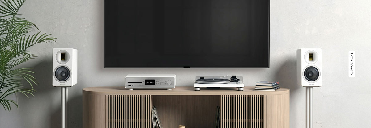 Zwei sonoenro ORCHESTRA Slim (Gen.2) in Weiß stehen auf Ständern vor einr weißen Wand, dazwischen eine helle Holzkommode mit einem sonoro MAESTRO in Weiß-Silber und einem sonoro PLATINUM Automatic EAS in Weiß-Silber, dahinter ein großer Fernseher an der Wand