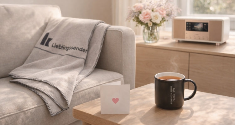 Blick ins Wohnzimmer, mit einem Sofa, wo die Klassik Radio Kuscheldecke liegt, auf dem Beistelltisch steht die Klassik Radio Tasse und im Hintergrund auf der Holzkommode ein Radio und Blumenstrauß