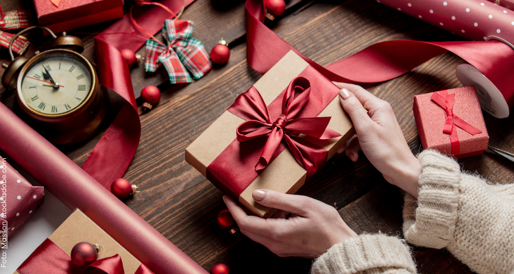 Frau hält ein fertig verpacktes Geschenk in der Hand. Das Geschenk ist mit goldenen Geschenkpapier eingepackt und drumerhum eine rote Schleife. Um das Geschänk und die Hände liegen noch Geschenkpapier, eine Uhr, weitere eingepackte Geschenke und Schleifen.
