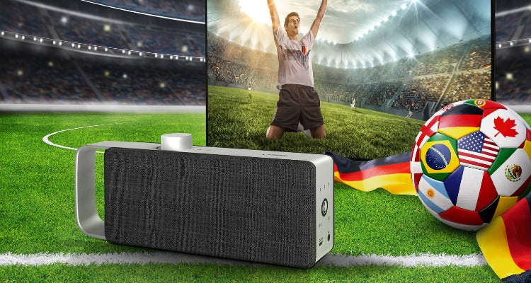 Fußballstation im Hinterrund, vorne ein Fernseher mit dem Bild eines jubelnden Fußballspielers und dem TV Sprachverstärker OSKAR, seitlich ein Ball mit den Flaggen der Nationen