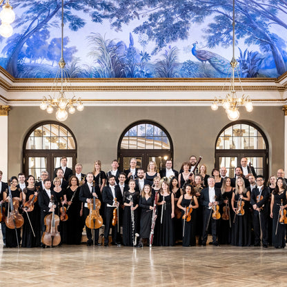 Totale des Münchner Symphoniker Orchesters in dunkler Kleidung, jeweils ihr Instrument in der Hand, stehend in einem orpulenten Saal mit blauer Deckenmalerei und Kronleuchtern an der Decke