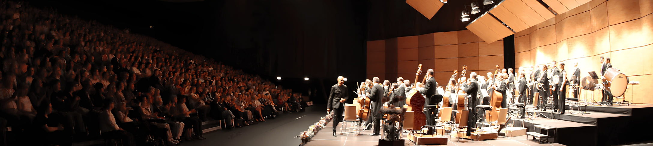 Panorama einer Supertotalen eines dunklen Konzertsaals, links volle Publikumsplätze, rechts eine beleuchtete Bühne mit einem klassischen Orchester