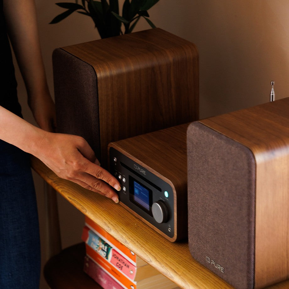 Person steht links neben PURE CLASSIC Stereo Mini in Coffee-Black, das auf einem Holzregal steht, und drückt einen Knopf am Radio