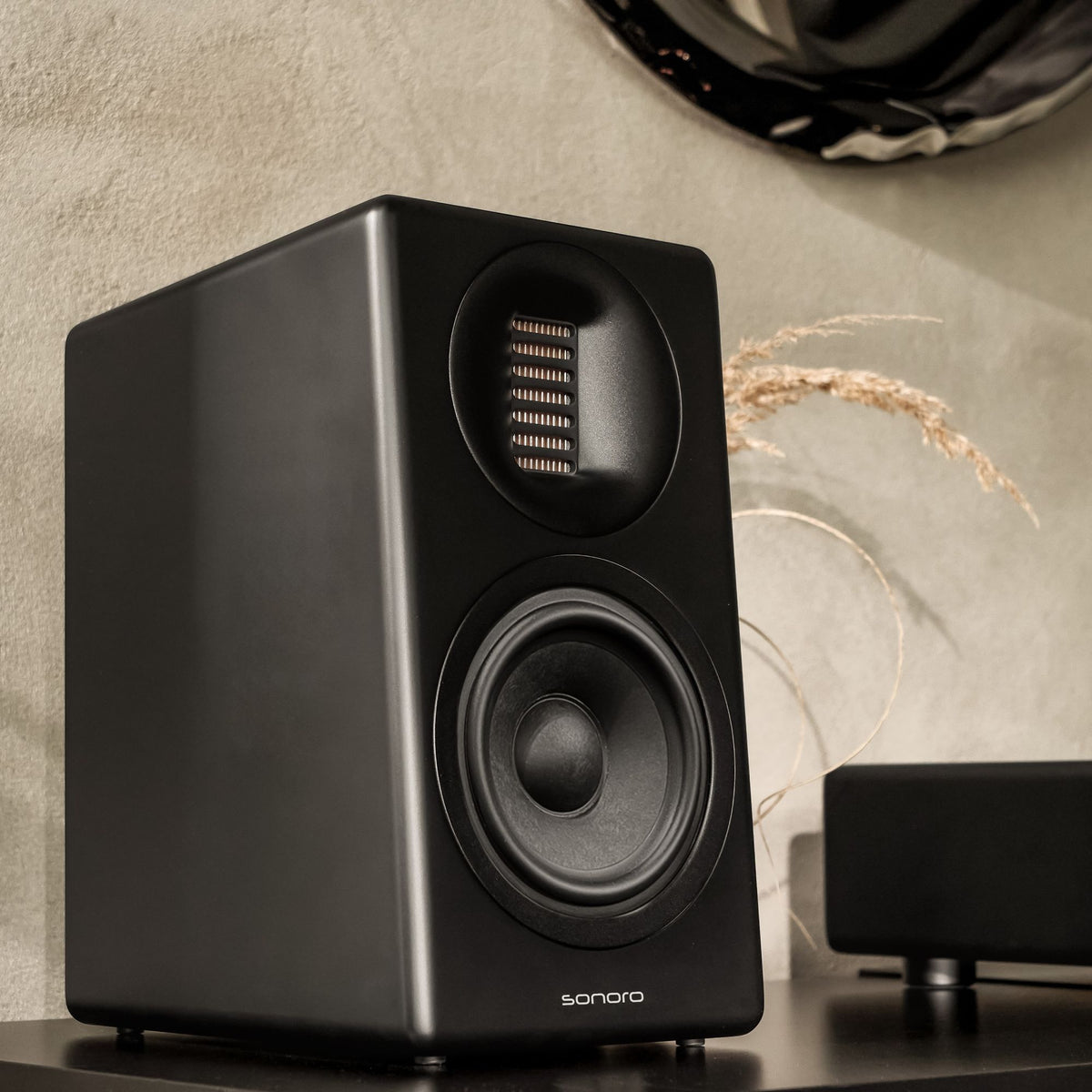 sonoro ORCHESTRA SLIM (Gen. 2) in Schwarz steht auf einer schwarzen Holzkommode vor einer cremefarbenen Wand, hinter dem Lautsprecher einige Getreideähren als Deko in einer Vase.